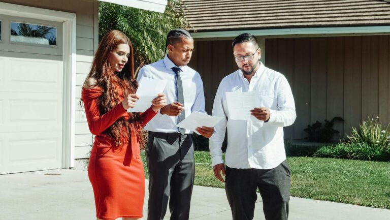 A group of adults reviewing real estate documents outside a house on a sunny day.